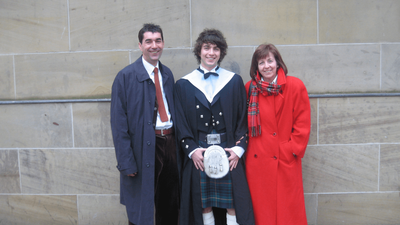 Jack wearing a kilt and graduation gown, standing between his smiling parents.